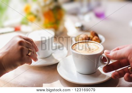 close-up, male and female hand holding a cup of coffee. couple in a cafe. focus on the crema, foam. light from a window. white cup of coffee on a saucer.