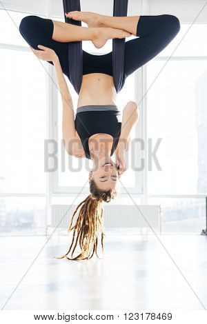 Cheerful young woman doing aerial yoga and talking on mobile phone in stusio