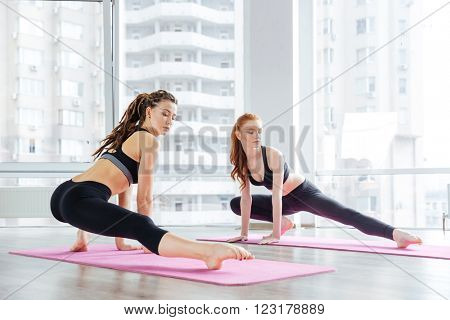 Two beautiful young women stretching legs together in yoga studio