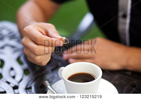 Close up shot of groom's hand is holding gold engagement ring with diamond and a cup of coffee on background