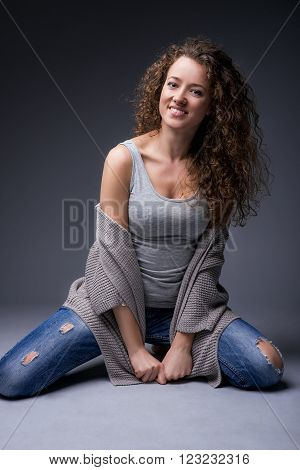 Attractive smiling girl in a studio on a black background. She looks at the camera with one hand down while the other hand on his head. She wears dark blue jeans and a gray sweater.