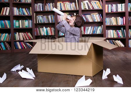 Playful childhood. Little boy having fun with cardboard box. Boy pretending to be pilot. Boy playing at room with bookshelf