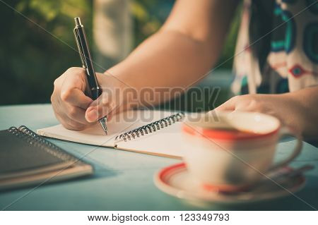 Woman hand writing journal on small notebook at outdoor area in cafe with morning scene and vintage filer effect