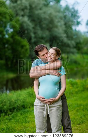 Happy smiling handsome man embracing and kissing his beautiful young pregnant woman who holding her belly in summer park. Happy family and pregnancy concept. Mother's Day