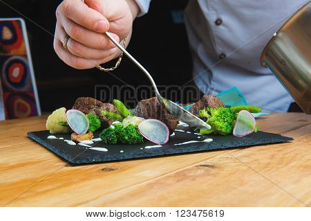 Professional chef decorate plate with meat greens. Man prepares delicious meals in kitchen. Chunks of roasted meat on  black plate with greens, radish, broccoli, asparagus and sauce. Close-up. cook pours sauce meal. In the hands of a spoon.