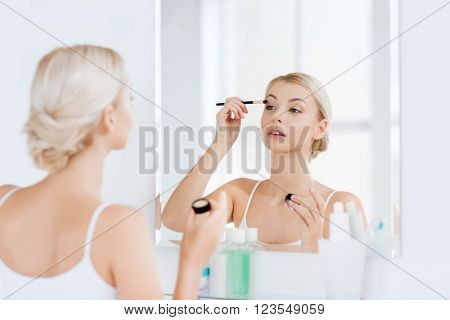 beauty, make up, cosmetics, morning and people concept - young woman applying eyeshade with makeup brush and looking to mirror at home bathroom