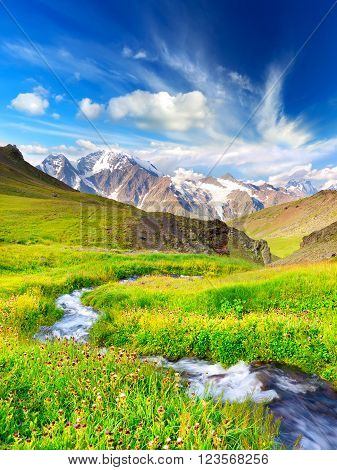 River on bright green meadow. High mountain and clouds.