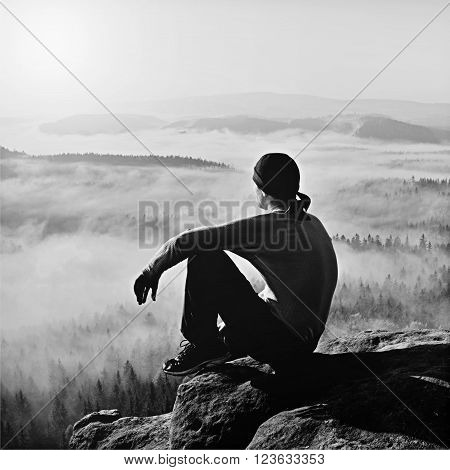 Man sit on the peak of rock and watching into colorful mist and fog in forest valley.