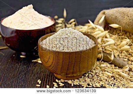 Bran flakes oat and oat flour in two wooden bowl, a bag of grain oats, oat ears against the dark wooden board