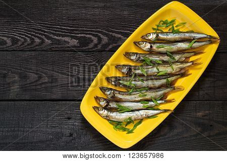 Smoked capelin with green onions on yellow plate on dark wooden background. Top view
