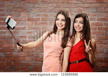 Two teenager girls making photo by their self with mobile phone on brick wall background