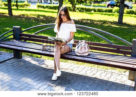 Beautiful girl student with glasses and a bag of coffee, sitting on a park bench, reading a book, fashion style, school life, in the summer on a sunny day in the fresh air. Woman relaxing.