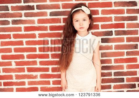 Cute kid girl 5-6 year old wearing style clothes over brick wall looking at camera. Childhood.