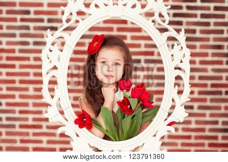 Smiling kid girl 5-6 year old holding red tulips in room over brick wall. Looking at camera. Posing in white vintage