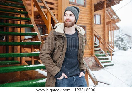 Pensive attractive bearded man standing near wooden cottage in snowy weather in winter