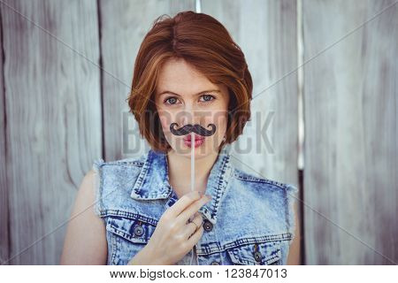 beautiful hipster woman holding a fake mustache to her face, against a wooden backhround