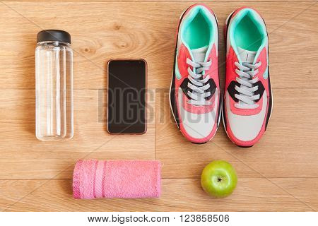 red and grey sneakers with grey shoelaces and red towel green apple bottle with water mobile phone with white headphones on wooden background indoors