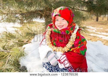 Child girl in Russian pavloposadskie folk scarf on head with floral print and with bunch of bagels on background of snow. Portrait of girl dressed in Russian style