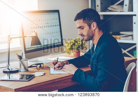 Working moments. Side view of confident young man writing something in his notebook while sitting at his working place in office

** Note: Visible grain at 100%, best at smaller sizes
