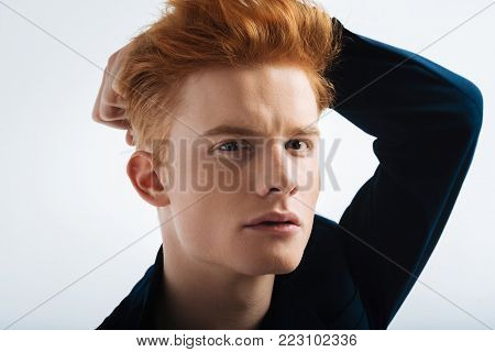 Handsome boy. Good-looking stony-faced red-headed young man wearing a black jacket and touching his head and staring