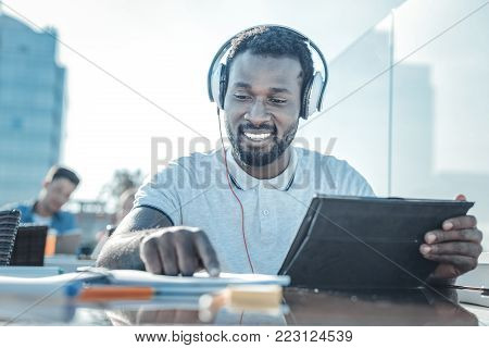 Education is the key to success. Low angle shot of a cheerful college guy smiling while enjoying the music playing in his headphones and working on a home assignment.