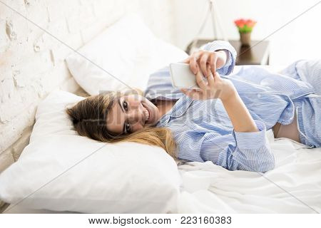 Pretty young flirty woman relaxing on her bed and using a smartphone to text her boyfriend