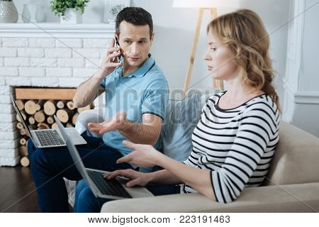 Freelancing. Handsome concentrated dark-haired man talking on the phone and working on his laptop and discussing work with his wife