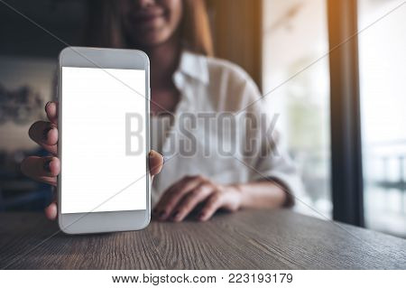 Mockup image of a woman holding and showing white mobile phone with blank screen on the table in modern cafe