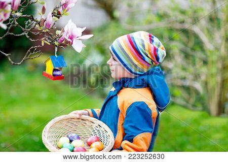 Cute adorable little kid boy making an egg hunt on Easter. Happy child searching and finding colorful eggs in domestic garden. Boy in spring clothes on cold day. Old christian and catholoc tradition