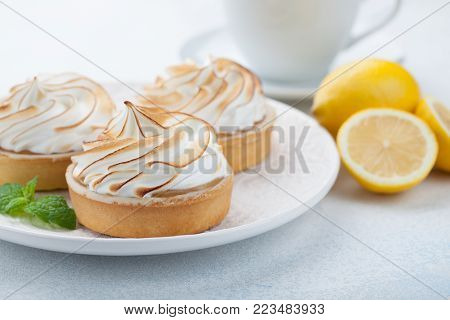 Lemon tartlets with meringue on vintage white plate and Cup of tea on the stone table. Tasty treat on a light blue background.