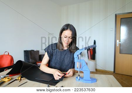 female fixing snap on leather pattern using special mechanism. Young designer decided to try to make women bags for sale. Black-haired girl with red nails wearing grey T-shirt sitting near table in workshop. Concept of creative work, own business or handw