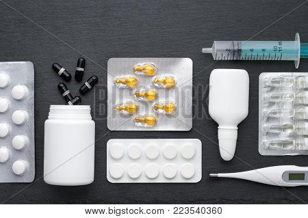 Thermometer, nasal drops tablets and ampoules with medicine on dark slate board. Top view, flat lay. Meds for flue treatment and antibiotics, health care concept