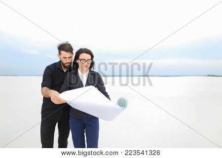 Deputy female and architect male meet to discuss stadium in countryside, people standing in deserted place holding whatman paper with plan smiling looking around. Young bearded man in black shirt and lady in glasses wearing strict suit communicating. Conc