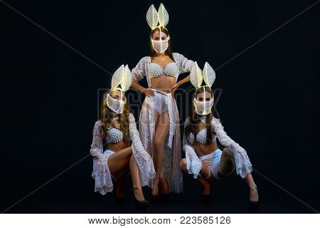 Group of Sexy girsl in white lingerie and LED masks. Go-go dancers. Three Strippers posing in the studio on a black background. Girls show ballet in the night club. show-program in a night club