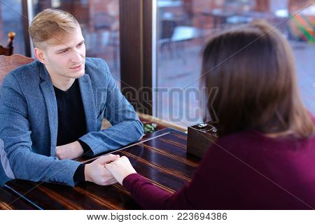 Boyfriend want to make girlfriend offer of hand and heart in favorite cafe, couple waiting for order and fooling. Young fair-haired woman with neat red manicure and guy in blue jacket sitting near wooden table. Concept of cozy luncheonette and nice atmosp