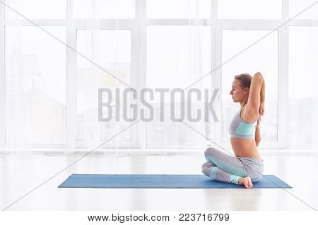 Beautiful young woman practices yoga asana Garudasana - Eagle pose at the bright yoga class with large windows