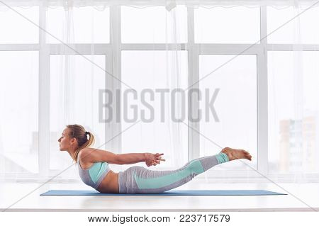 Beautiful woman practices yoga asana Salabhasana - locust pose at the bright yoga class with large windows