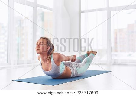 Beautiful woman practices yoga asana Salabhasana - locust pose at the bright yoga class with large windows