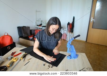 female fixing snap on leather pattern using special mechanism. Young designer decided to try to make women bags for sale. Black-haired girl with red nails wearing grey T-shirt sitting near table in workshop. Concept of creative work, own business or handw