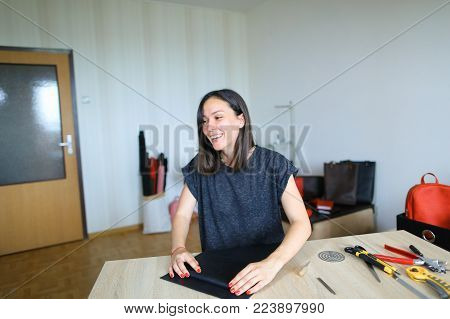 female fixing snap on leather pattern using special mechanism. Young designer decided to try to make women bags for sale. Black-haired girl with red nails wearing grey T-shirt sitting near table in workshop. Concept of creative work, own business or handw