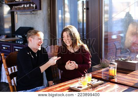 Couple sitting near table with sea-buckthorn broth in glass cups jug cutlery in cafe discuss film, young people go to favorite luncheonette after movie show. Handsome male in black T-shirt and Asian girl in burgundy dress holding hands and sharing impress