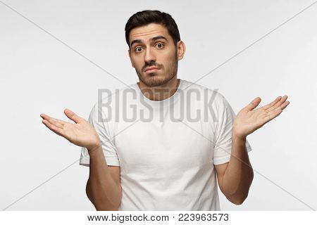 I don't know. Young man isolated on grey background being at a loss, showing helpless gesture with arm and hands, mouth curved as if he does not know what to do