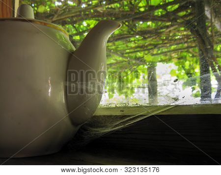 An Old White Ceramic Teapot Stands In Pownig On A Windowsill. The Empty House Was Covered With Dust 