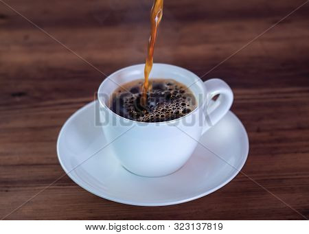 Coffee Pouring Down In White Cup, Close Up View. White Coffee Cup And Saucer On Brown Wooden Table. 
