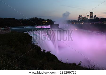 Wide view of Niagara Falls illuminated pink in evening as seen from USA, with water flow all blurred, cityscape visible on Canada side