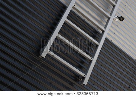 Close-up Of Metal Ladder And Surveillance Videocamera Against Junction Of Black And White Corrugated