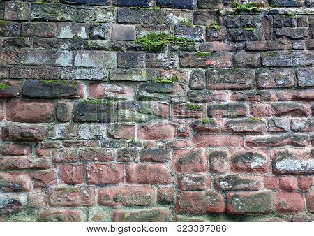 Full Frame Image Of An Ancient Sandstone Wall In Red And Black Colors With Sodd And Algae Growing On