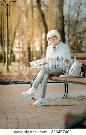 Pleasant Superannuated Lady In Glasses Thoughtfully Reading Newspaper On Bench