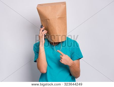 Teen boy with paper bag over head speaking on mobile phone, isolated on white background. Teenager cover head with shopping bag holds smart phone. Child pulling bag over head.