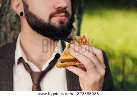 Young Attractive Business Man With Beard Sitting On Green Grass Under Tree And Resting In Park. Bite
